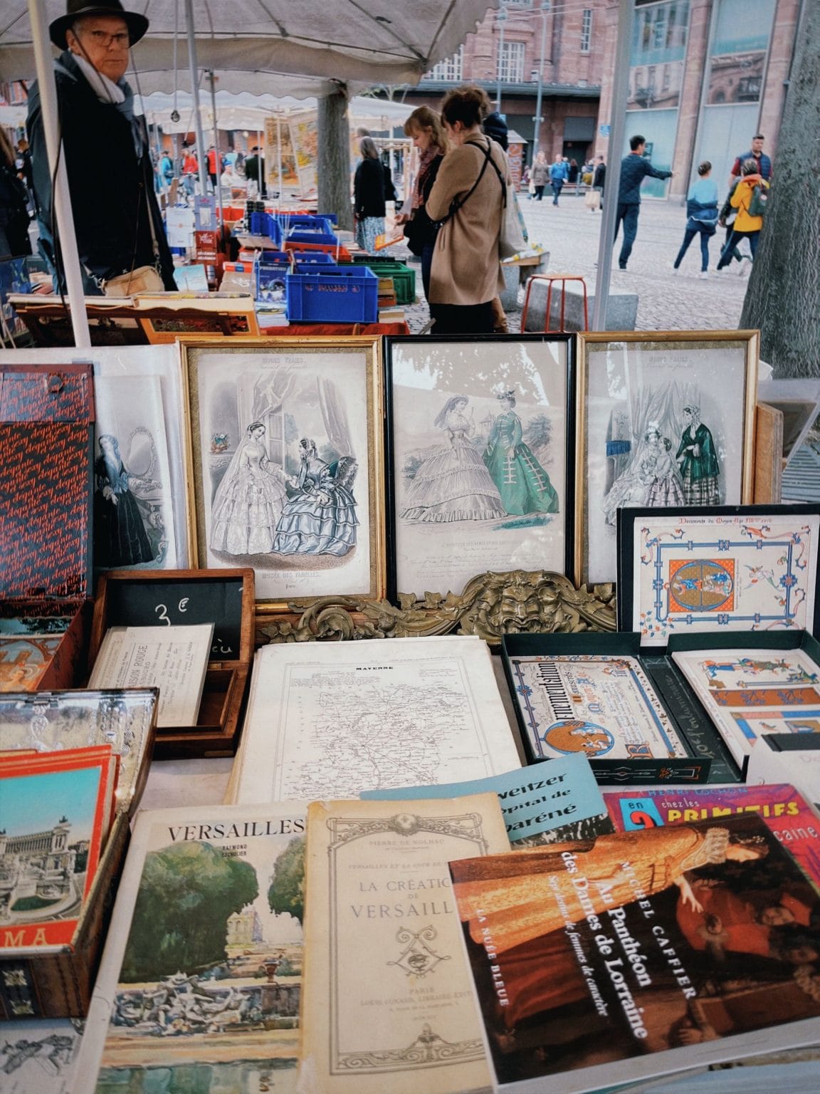 Étal de brocante sous un chapiteau : gravures anciennes encadrées, vieux livres et cartes posés sur une table, avec une ardoise à 3 euros