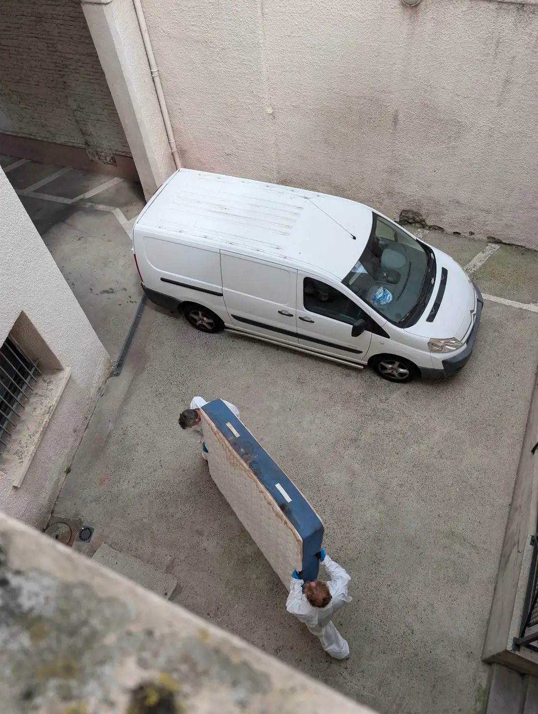 Vue plongeante sur la cour d'un immeuble : deux intervenants de Soli'Débarras en combinaison portent un matelas jusqu'à l'utilitaire