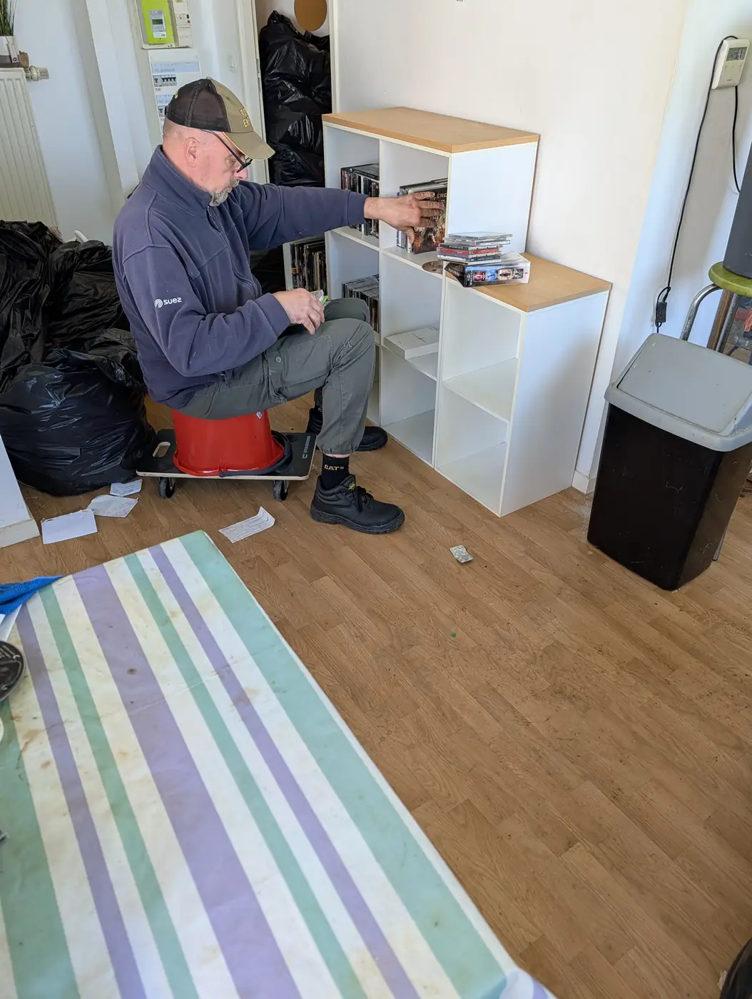 Intervenant assis, examinant un à un les DVD d'une étagère avant évacuation, dans un appartement en cours de débarras