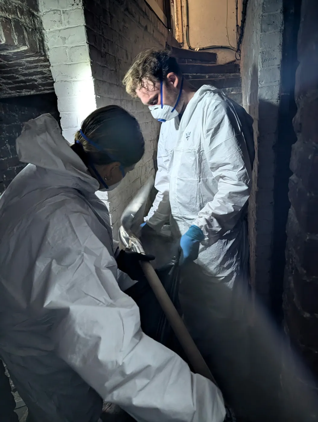 Deux intervenants de Soli'Débarras en combinaison et masque, évacuant les déchets à la pelle dans un sous-sol