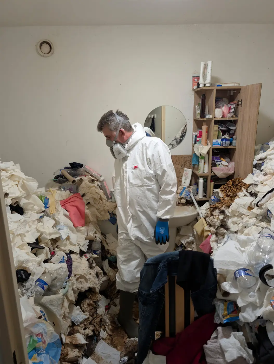 Intervenant de Soli'Débarras en combinaison, masque respiratoire et gants, dans une salle de bain entièrement recouverte de déchets