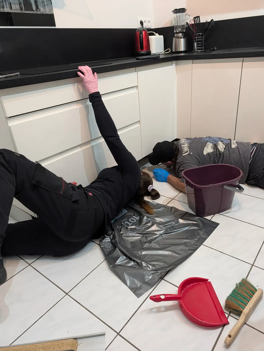 Deux intervenants de Soli'Débarras allongés au sol, nettoyant sous les meubles d'une cuisine
