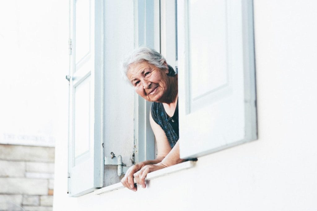 Femme âgée souriante accoudée à la fenêtre de son logement, volets clairs ouverts